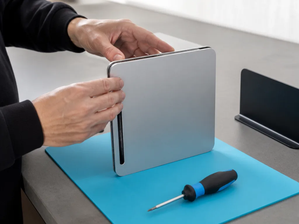 Photorealistic editorial photograph: hands adjusting a plain brushed-aluminium console on a grey workbench, screwdriver
